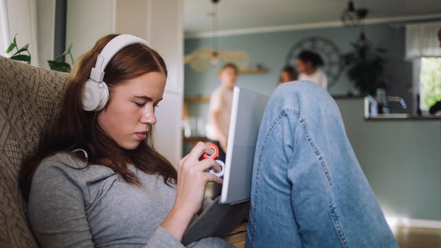 Eine junge Frau liegt halb aufgerichtet auf einem Sofa. Sie trägt weiße Kopfhörer und schaut konzentriert auf ein Tablet, das auf ihrem Bauch steht. In ihren Händen hält sie ein so genanntes Fidget Toy, das der Entspannung dienen soll. Im Hintergrund sind unscharf zwei Personen in einer Küche zu sehen.