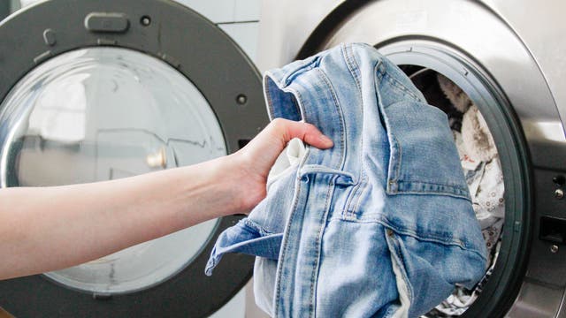Eine Person legt eine blaue Jeans in eine geöffnete Waschmaschine. Die Waschmaschine ist aus Edelstahl und befindet sich in einem modernen Waschraum. Im Hintergrund ist eine weitere Waschmaschine zu sehen.