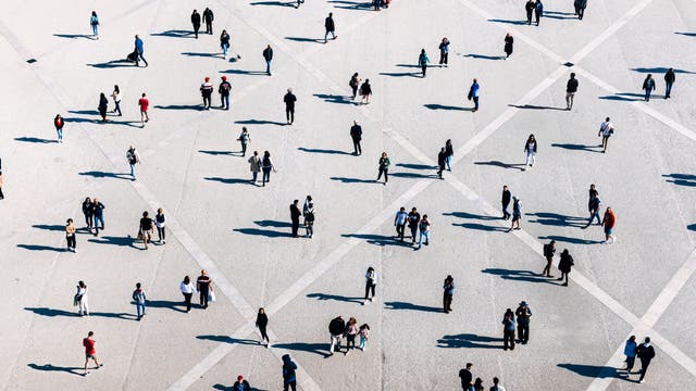 Eine große, offene Fläche mit vielen Menschen, die in kleinen Gruppen oder allein stehen und gehen. Die Szene ist aus der Vogelperspektive aufgenommen, wodurch geometrische Muster auf dem Boden sichtbar werden. Die Personen werfen lange Schatten. Die Kleidung der Menschen ist vielfältig, was auf eine alltägliche, urbane Umgebung schließen lässt.