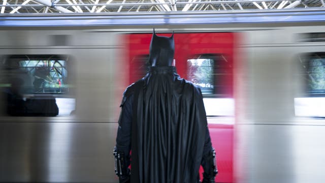 Eine Person in einem Batman-Kostüm steht auf einem Bahnsteig und blickt auf einen vorbeifahrenden Zug. Der Zug ist in Bewegung, was durch die verschwommenen Fenster sichtbar ist. Die Szene spielt sich in einer modernen, überdachten Bahnhofsumgebung ab.