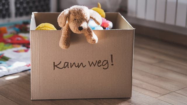 Ein Pappkarton steht auf einem Holzboden. Auf dem Karton steht in handgeschriebener Schrift "Kann weg!". Ein Plüschtier in Form eines Hundes hängt über den Rand des Kartons und schaut in die Kamera. Im Hintergrund links ist ein bunter Kinderteppich zu sehen.
