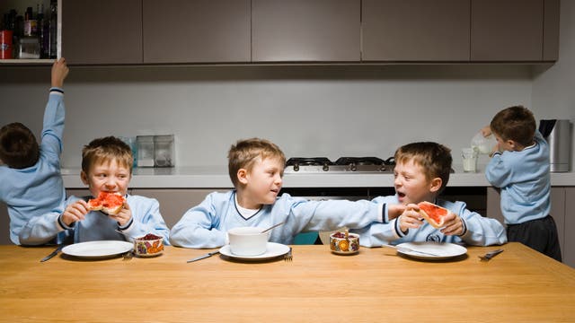 Ein Junge in einem blauen Pullover sitzt an einem Tisch und isst ein Marmeladenbrot. Neben ihm sitzt ein weiterer Junge, der ihm das Brot wegnehmen möchte. Links von ihm isst ein weiterer Junge ebenfalls ein Marmeladenbrot. Auf dem Tisch stehen eine Schüssel, Tassen und Besteck. Im Hintergrund öffnet ein Junge einen Schrank, während ein anderer Milch in ein Glas gießt. 