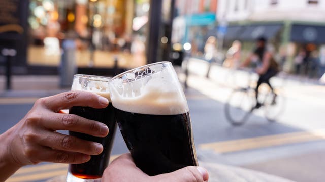 Zwei Personen stoßen im Freien bei gutem Wetter mit einem Guinness-Bier an, im Hintergrund sind Geschäfte und Passanten in Unschärfe zu sehen.