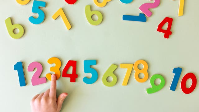 Eine Kinderhand zeigt auf bunte, magnetische Zahlen auf einer hellgrünen Oberfläche. Die Zahlen sind in verschiedenen Farben wie Rot, Blau, Gelb und Grün dargestellt und reichen von 1 bis 10. Die Anordnung der Zahlen ist spielerisch und unregelmäßig.