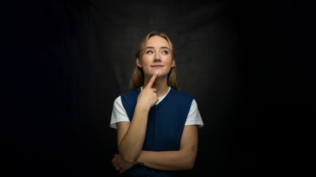 Eine Frau steht vor einem dunklen Hintergrund und schaut nachdenklich nach oben, während sie mit einem Finger an ihr Kinn tippt. Sie trägt ein blaues Oberteil über einem weißen T-Shirt. Die Szene vermittelt einen Moment des Nachdenkens oder der Überlegung.
