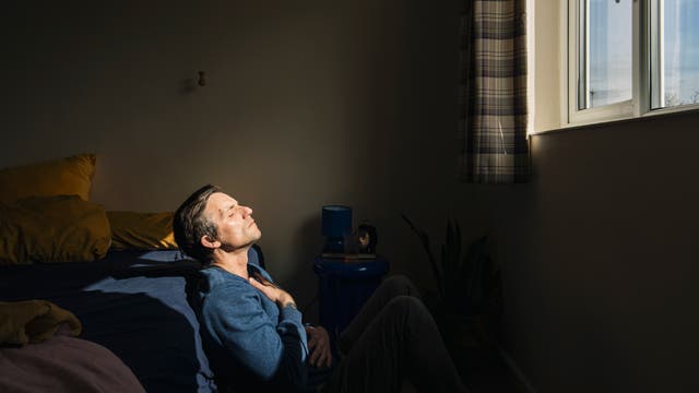 Eine Person sitzt auf dem Boden eines Schlafzimmers, lehnt sich an das Bett und genießt das Sonnenlicht, das durch ein Fenster fällt. Die Person trägt einen blauen Pullover und hat die Augen geschlossen, während sie eine Hand auf die Brust legt. Neben dem Bett steht ein kleiner Tisch mit einer Lampe und Büchern. Die Atmosphäre wirkt ruhig und entspannt.