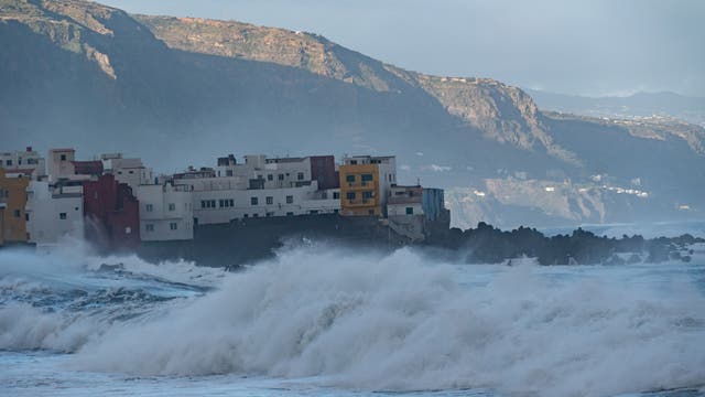 Küstenlandschaft mit stürmischer See und hohen Wellen, die gegen eine Reihe bunter Gebäude schlagen. Im Hintergrund sind felsige Klippen zu sehen, die von der untergehenden Sonne beleuchtet werden. 