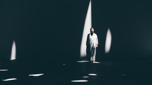 Eine Frau steht in einem großen, dunklen Raum, beleuchtet von schmalen Lichtstrahlen, die durch die Decke fallen. Sie trägt lange, helle Kleidung und hat den Kopf mit geschlossenen Augen leicht nach hinten gelegt, wie wenn sie das Licht genießt. Die Szene könnte Teil einer Theateraufführung sein.