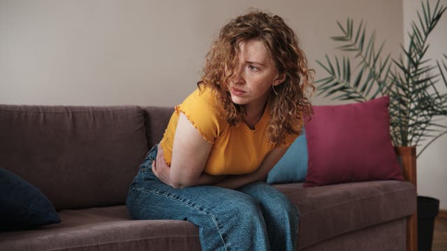 Eine Frau mit lockigem Haar sitzt auf einem Sofa und hält sich den Bauch, als ob sie Schmerzen hätte. Sie trägt ein gelbes T-Shirt und blaue Jeans. Im Hintergrund sind bunte Kissen und eine Zimmerpflanze zu sehen.