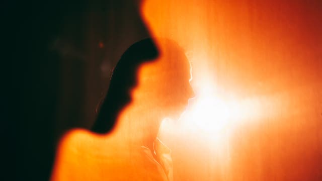 Eine Silhouette einer Person vor einem leuchtend orangefarbenen Hintergrund. Die Person ist seitlich abgebildet, und das Licht erzeugt einen starken Kontrast, der die Konturen betont. Die Szene wirkt künstlerisch und atmosphärisch.