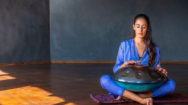 Eine Person sitzt in einem ruhigen Raum auf einem Teppich und spielt ein Handpan-Instrument. Sie trägt ein blaues Outfit und hat die Augen geschlossen, während sie das Instrument mit den Händen berührt. Der Raum ist spärlich beleuchtet, mit einem dunklen, strukturierten Hintergrund. Die Szene vermittelt eine Atmosphäre der Entspannung und Konzentration.