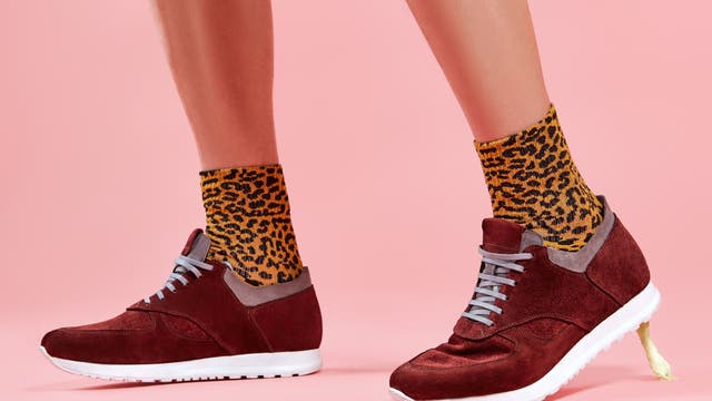 Ein Paar Beine in roten Turnschuhen mit grauen Schnürsenkeln und orangefarbenen Socken mit Leopardenmuster auf einem rosa Hintergrund. Der hintere Schuh klebt an der Ferse durch ein Kaugummi am Boden fest.