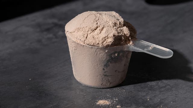 Ein transparenter Messlöffel, gefüllt mit beigem Pulver, steht auf einer dunklen, strukturierten Oberfläche. Das Pulver ist leicht über den Rand des Löffels gehäuft. Einige Pulverreste sind auf der Oberfläche verstreut. Das Bild zeigt eine Nahaufnahme, die die Textur des Pulvers betont.