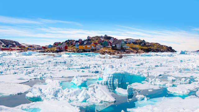 Bunte Häuser auf einem felsigen Hügel in einem grönländischen Dorf, umgeben von einer weiten, eisbedeckten Landschaft. Im Vordergrund sind große Eisblöcke und schimmerndes blaues Wasser zu sehen. Der Himmel ist klar und blau, was die kühle, ruhige Atmosphäre der arktischen Umgebung unterstreicht.