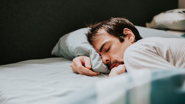 Eine Person liegt schlafend auf der Seite in einem Bett mit hellblauer Bettwäsche. Der Kopf ruht auf einem Kissen, und die Person trägt ein gestreiftes Oberteil. Im Hintergrund ist ein Teil des Bettrahmens und ein weiteres Kissen zu sehen.