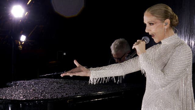 Celine Dion in einem glitzernden, weißen Kleid steht auf dem Eiffelturm und singt in ein Mikrofon. Ihre rechte Hand ist ausgestreckt, während sie performt. Im Hintergrund ist ein Pianist zu sehen, der auf einem schwarzen Flügel spielt. Die Umgebung erscheint dunkel, mit einigen hellen Lichtern im Hintergrund.