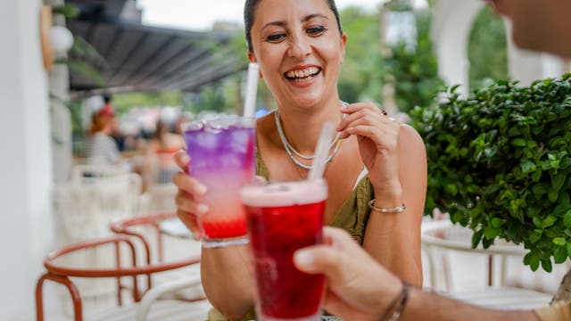 Eine Person sitzt in einem Café im Freien und hält ein lila Getränk mit einem Strohhalm. Sie lächelt und stößt mit einer anderen Person an, die ein rotes Getränk hält. Im Hintergrund sind unscharf weitere Gäste und Pflanzen zu sehen. Die Szene wirkt entspannt und gesellig.