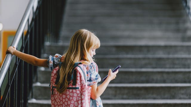 Ein Kind mit einem rosa Rucksack steht auf einer Treppe und schaut auf ein Smartphone. Es hält sich mit einer Hand am Geländer fest. Die Umgebung wirkt wie ein Schulgebäude.