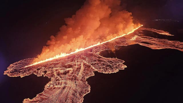 Luftaufnahme eines Vulkanausbruchs bei Nacht. Eine lange, glühende Lavafront zieht sich diagonal durch das Bild, umgeben von dichten Rauchwolken, die in den Nachthimmel aufsteigen. Die Lava leuchtet intensiv orange und rot, während sie sich über die dunkle Landschaft ausbreitet. Im Hintergrund sind schwach beleuchtete Stadtlichter zu erkennen.