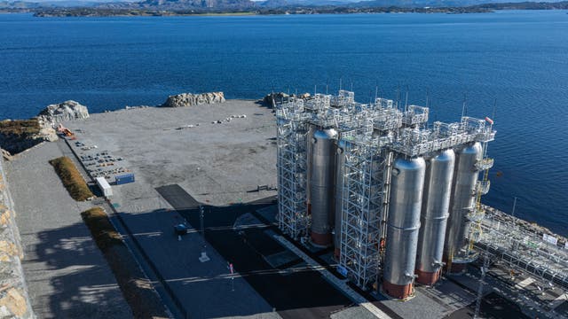 Industrieanlage an der Küste mit mehreren großen, silbernen Tanks und Rohrleitungen. Die Anlage befindet sich auf einem weitläufigen, gepflasterten Gelände neben einem tiefblauen Gewässer. Im Hintergrund sind Hügel und eine bewaldete Landschaft zu sehen. Der Himmel ist klar mit wenigen Wolken.