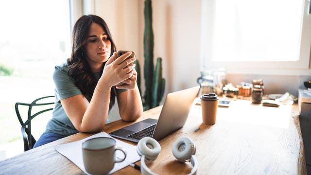 Eine Person sitzt an einem Holztisch und schaut auf ihr Smartphone. Auf dem Tisch befinden sich ein Laptop, eine Tasse, Kopfhörer und ein Kaffeebecher. Im Hintergrund steht ein Kaktus neben einem Fenster, durch das Tageslicht hereinfällt. Die Szene vermittelt eine entspannte Arbeits- oder Lernumgebung zu Hause.
