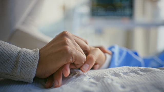 Eine Person hält die Hand einer anderen Person, die im Bett liegt. Im Hintergrund ist unscharf medizinisches Equipment zu erkennen. Die Szene vermittelt Fürsorge und Unterstützung in einer klinischen Umgebung.