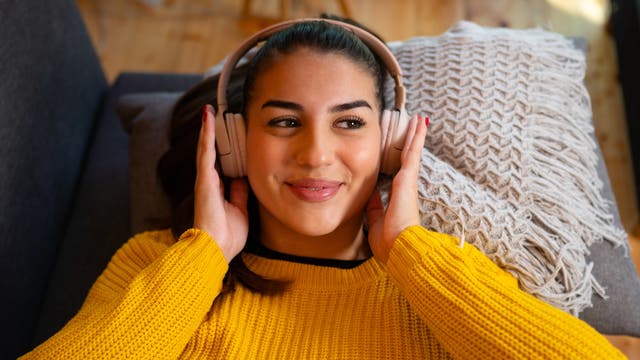 Eine Frau liegt entspannt auf einem Sofa und hört Musik mit großen Kopfhörern. Sie trägt einen gelben Pullover und lächelt leicht. Neben ihr liegt ein dekoratives Kissen mit Fransen. Der Raum hat einen Holzboden und wirkt gemütlich.