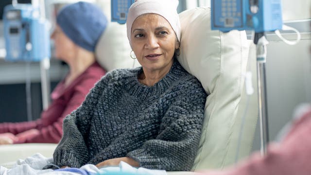 Zwei Frauen, die vordere mit einer hellen Mütze, die hintere mit einer blauen Mütze, sitzen auf Behandlungsstühlen in einer onkologischen Klinik.