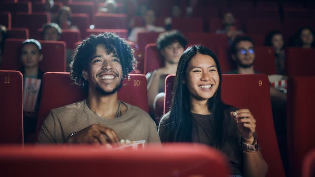 Eine Gruppe von Menschen sitzt in einem Kino auf roten Sitzen, die mit Nummern versehen sind. Zwei Personen im Vordergrund lächeln und halten Popcorn, während sie den Film genießen. Die Atmosphäre ist entspannt und fröhlich, mit gedämpftem Licht im Hintergrund.