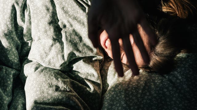 Eine Person liegt in einem Bett mit gemusterter Bettwäsche und hält eine Hand schützend vor das Gesicht, während Sonnenlicht auf sie fällt. Die Szene vermittelt ein Gefühl von Morgenruhe und Geborgenheit.