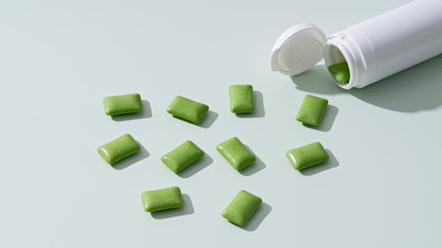 Eine weiße Plastikdose liegt auf einer hellen Oberfläche, aus der mehrere grüne Kaugummis herausgefallen sind. Die Kaugummis sind rechteckig und glänzend. Die Dose ist geöffnet und zeigt einen weiteren Kaugummi im Inneren. Die Szene ist minimalistisch und sauber gestaltet.