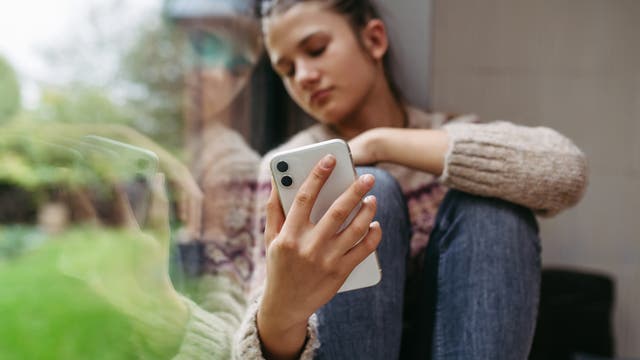 Eine Person sitzt nachdenklich an einem Fenster und hält ein Smartphone in der Hand. Ihr Blick ist auf das Gerät gerichtet, während ihr Spiegelbild im Glas sichtbar ist. Sie trägt einen Strickpullover und Jeans. Draußen ist unscharf eine grüne Landschaft zu erkennen.