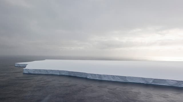 Ein großer, flacher Eisberg schwimmt im offenen Meer unter einem bewölkten Himmel. Die Oberfläche des Eisbergs ist glatt und weiß, während die Seiten steil ins Wasser abfallen. Das Meer ist dunkel und ruhig, und der Himmel zeigt diffuse Sonnenstrahlen, die durch die Wolken brechen.