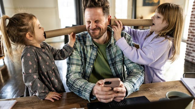 Ein Mann sitzt an einem Tisch mit einem Laptop und einem Smartphone in der Hand. Zwei Kinder stehen neben ihm und halten ein Papprohr an seine Ohren, während sie hineinsprechen. Der Mann kneift die Augen zusammen und lächelt leicht. Auf dem Tisch liegen Notizbücher und eine Tasse. Die Szene spielt in einem hellen Raum mit Fenstern im Hintergrund.