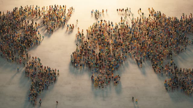 Eine große Menschenmenge bildet die Umrisse einer Weltkarte auf einem hellen Hintergrund. Die Personen sind in verschiedenen Farben gekleidet und stehen dicht beieinander, um die Kontinente darzustellen. Die Szene wird von oben betrachtet und zeigt die Vielfalt und Einheit der Menschheit.