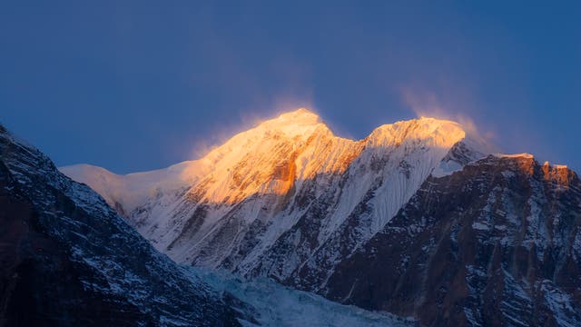 Ein schneebedeckter Berggipfel wird von der aufgehenden Sonne in warmes, goldenes Licht getaucht. Der Himmel ist klar und tiefblau, während die Sonnenstrahlen die schroffen Felswände und die Schneedecke des Berges hervorheben. Die Szene vermittelt eine ruhige und majestätische Atmosphäre.