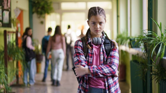 Ein junges Mädchen mit Kopfhörern um den Hals steht mit verschränkten Armen in einem Schulflur. Sie trägt ein kariertes Hemd und schaut ernst. Im Hintergrund sind unscharf andere Schüler zu sehen, die den Flur entlanggehen. Der Flur ist mit Pflanzen dekoriert und hat grüne Wände.