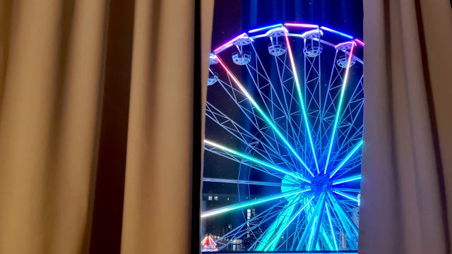 Ein beleuchtetes Riesenrad mit bunten Lichtern ist durch einen Spalt zwischen zwei Vorhängen sichtbar. Die Szene spielt sich nachts ab, und die Lichter des Riesenrads leuchten in verschiedenen Farben, darunter Blau, Grün und Rot. Im Hintergrund sind schwach beleuchtete Gebäude zu erkennen.