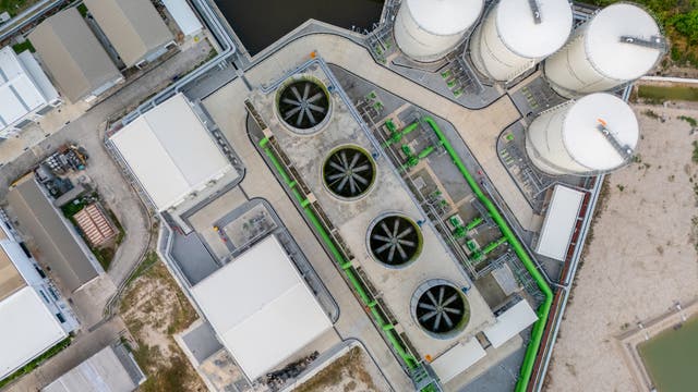 Luftaufnahme einer industriellen Anlage mit mehreren großen, runden Silos und rechteckigen Gebäuden. In der Mitte befinden sich vier große Ventilatoren, die Teil eines Kühlsystems zu sein scheinen. Die Anlage ist von Straßen und Vegetation umgeben. Die Struktur und Anordnung der Gebäude und Silos deuten auf eine komplexe industrielle Nutzung hin.