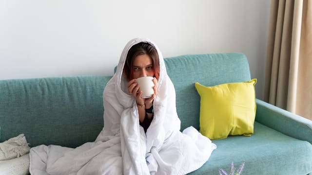 Eine Person sitzt auf einem türkisfarbenen Sofa, in eine weiße Decke gehüllt, und hält eine Tasse. Neben ihr liegt ein gelbes Kissen. Im Vordergrund ist ein Tisch mit einer Pflanze zu sehen. Die Szene vermittelt ein Gefühl von Gemütlichkeit und Entspannung.