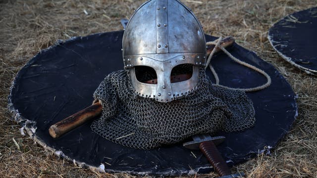 Ein mittelalterlicher Helm aus Metall liegt auf einem runden Schild, umgeben von einer Kettenhaube. Daneben befinden sich ein Holzgriff und ein Schwert. Der Hintergrund besteht aus trockenem Gras.