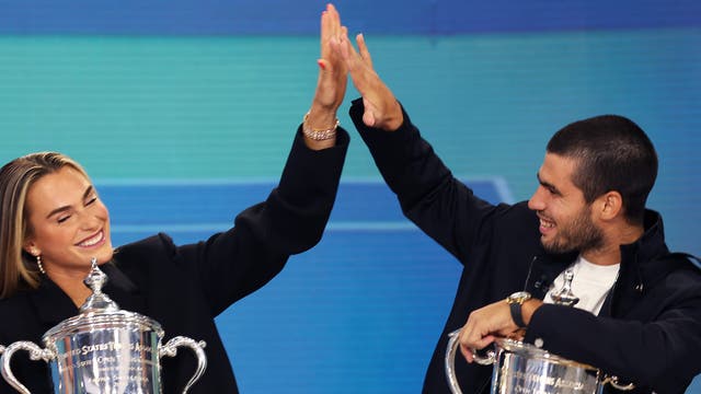 Zwei Personen sitzen nebeneinander und geben sich ein High-Five. Beide halten jeweils einen großen silbernen Pokal in der anderen Hand. Die Szene vermittelt Freude und Erfolg. Der Hintergrund ist blau.