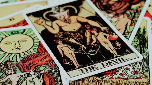 Ein Stapel Tarotkarten, im Vordergrund die Karte "The Devil". Die Karte zeigt eine mythologische Figur mit Hörnern, die über zwei angeketteten Menschen thront. Um die Hauptkarte herum sind weitere Tarotkarten mit verschiedenen Symbolen und Illustrationen sichtbar, darunter eine Karte mit einer Sonne. Die Farben sind lebendig und kontrastreich, was den mystischen und symbolischen Charakter der Karten betont.