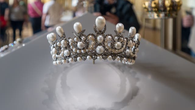 Eine mit Perlen und Diamanten verzierte Tiara wird in einem Museum ausgestellt. Im Hintergrund sind unscharf mehrere Personen zu sehen, die die Ausstellung betrachten. Die Tiara ist auf einem weißen Podest platziert, und ihr Schatten ist deutlich sichtbar. Die Umgebung wirkt elegant und historisch.