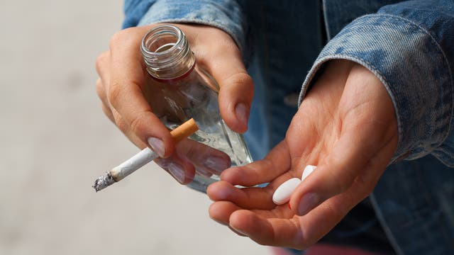 Eine Person in einer Jeansjacke hält in der einen Hand eine brennende Zigarette und eine leere Glasflasche, in der anderen Hand zwei weiße Tabletten. Die Szene vermittelt den Eindruck von Substanzmissbrauch.
