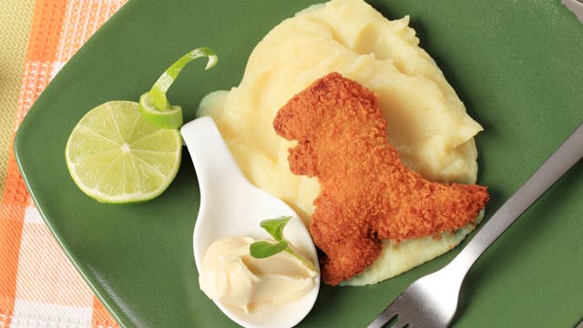 Ein grüner Teller mit Kartoffelpüree, darauf ein paniertes Stück Fleisch in Form eines Dinosauriers. Daneben eine weiße Löffelschale mit einem Klecks Butter und einem kleinen grünen Blatt. Eine halbe Limettenscheibe liegt am Rand. Eine Gabel ist am rechten Rand des Tellers platziert. Der Hintergrund zeigt ein orange-weiß kariertes Tischtuch.