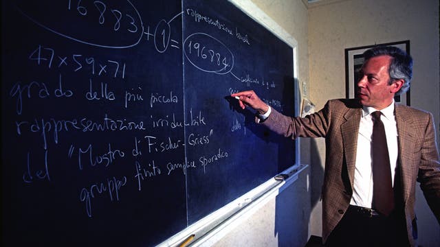 Ein Mann in einem Anzug steht vor einer Tafel und zeigt auf eine mathematische Formel. Die Formel ist in Kreisen hervorgehoben. Der Mann scheint die mathematischen Konzepte zu erklären. Der Raum ist schlicht eingerichtet.