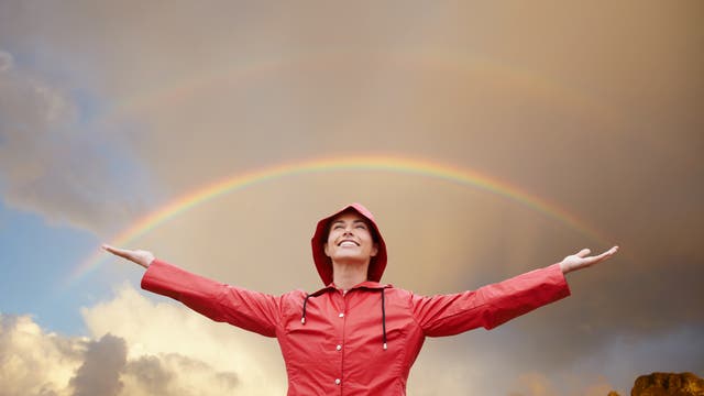 Eine Person in einem roten Regenmantel steht mit ausgebreiteten Armen vor einem doppelten Regenbogen am Himmel. Die Person lächelt und blickt nach oben. Im Hintergrund sind dramatische Wolken und ein Berg zu sehen. Die Szene vermittelt ein Gefühl von Freude und Naturverbundenheit.