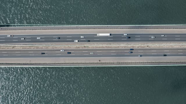 Luftaufnahme direkt von oben einer mehrspurigen Autobahnbrücke über einen Fluss. Mehrere Fahrzeuge, darunter Autos und ein Lastwagen, fahren in beide Richtungen. Die Brücke verläuft horizontal durch das Bild, während das Wasser des Flusses darunter sichtbar ist.