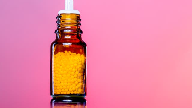 Eine braune Glasflasche mit einem weißen Verschluss, gefüllt mit homöopathischen Kügelchen. Der Hintergrund ist in einem sanften Rosa gehalten.